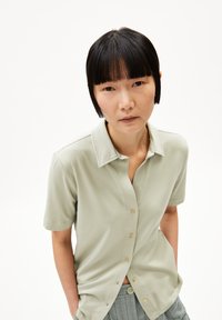 Camicia verde chiaro con colletto e maniche corte, dotata di chiusura con bottoni, realizzata in tessuto morbido con una consistenza liscia e senza fantasie.