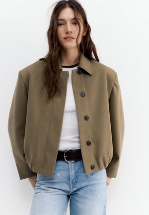 Hosszú barna hajú nő olívazöld gombos kabátban, fehér ingben, fekete övvel és világoskék farmerral, fehér háttér előtt.