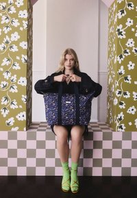 Ung kvinna som sitter mellan två blomsterpelare, håller en stor mörkblå blommig tygväska och har på sig gröna strumpor med bruna sandaler.