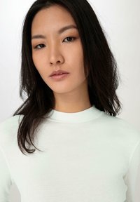 Langärmliges, hellminzfarbenes Oberteil aus weichem Stoff, mit hohem Kragen und glatter Textur. Das Modell hat welliges, dunkles Haar.