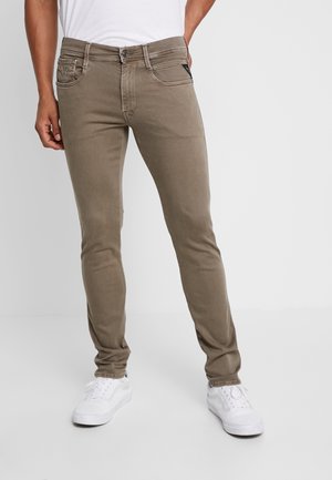 Man wearing slim fit taupe jeans with five pockets and white sneakers, standing with one hand in pocket on plain background.