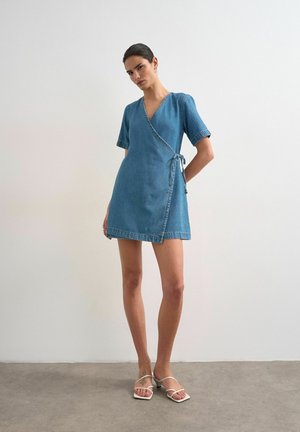 Vrouw staat tegen een effen muur, draagt een korte mouwen blauwe denim wikkeljurk, gestrikt in de taille, en witte sandalen met bandjes en hakken.