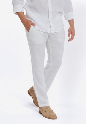 Uomo che indossa pantaloni di lino bianchi, camicia bianca abbottonata con maniche arrotolate e mocassini in camoscio beige, in piedi con una mano in tasca.