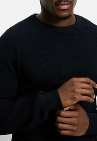 Marineblauer Strickpullover mit strukturiertem Muster, gerippten Bündchen und rundem Halsausschnitt. Nahaufnahme von Händen in entspannter Position.