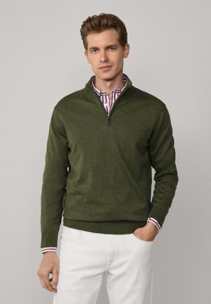 Dunkelgrüner Quarter-Zip-Pullover mit weicher Textur und gerippten Bündchen, über einem gestreiften Kragenhemd getragen, kombiniert mit weißen Hosen.