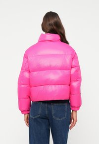 Veste rembourrée en rose vif, présentant un col haut, une coupe carrée, des poignets élastiques et une texture matelassée. Association avec un jean bleu foncé.