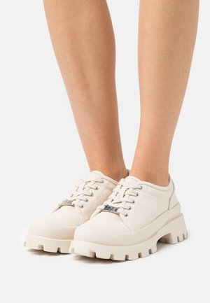 Zapatillas de lona blancas con suelas gruesas y texturizadas, y detalles de hardware plateado. Cuentan con un diseño de cordones y un cuello acolchado para mayor comodidad.