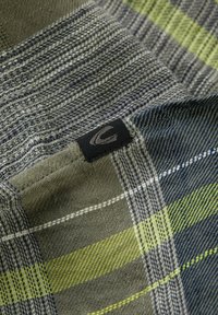 Vævet stof med en blanding af grønne, grå og blå ternede mønstre. Indeholder et sort mærke med et logo syet ind i stoffet.