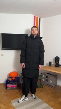 Dlhá čierna zimná bunda s vysokým golierom, vybavená predným zipsom a nenápadným logom. Nosí sa s čiernymi nohavicami a sivými teniskami na drevenej podlahe.