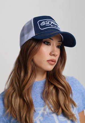 Joven mujer con cabello largo y castaño, que lleva una gorra Ed Hardy azul y blanca y un suéter azul claro, mirando hacia un lado.