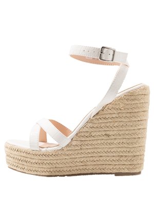 Biała sandałka na platformie i koturnie z teksturowanym, błyszczącym paskiem oraz regulowanym zapięciem wokół kostki. Koturn owinięty jutą dla dodatkowej wysokości i stabilności.