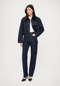 Giacca di jeans blu scuro con colletto in pelliccia sintetica, abbinata a jeans a gamba dritta dello stesso colore. Camicia bianca sotto, tacchi neri con punta a stiletto.