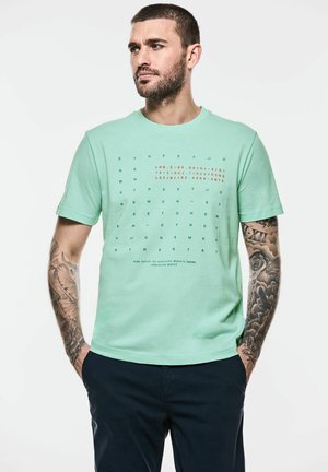 T-shirt en coton vert menthe avec des motifs de texte multicolores, manches courtes, col rond et coupe décontractée. Associé à un pantalon sombre.