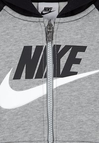 Hoodie zippé gris avec des accents noirs. Présente un logo Nike noir et blanc bien visible, une texture de tissu lisse et des détails de fermeture éclair argentés.