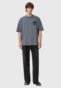 T-shirt cinza oversized com uma impressão gráfica preta no lado esquerdo do peito, combinada com jeans pretos de perna reta e calçado escuro.