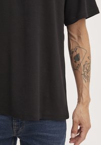 Camiseta negra de manga corta con un corte relajado. La tela parece suave y con textura. En el antebrazo se ve un tatuaje visible de cartas de juego.