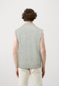 Pull sans manches gris clair, confectionné en tissu tricoté doux, avec un col rond et une bordure côtelée, porté sur un pantalon de couleur claire.
