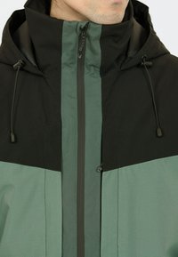Schwarze und teal-farbene Kapuzenjacke aus Stoff, ausgestattet mit einem Reißverschluss vorne, verstellbaren Kordeln und einer strukturierten Oberfläche mit einem Farbblockdesign.