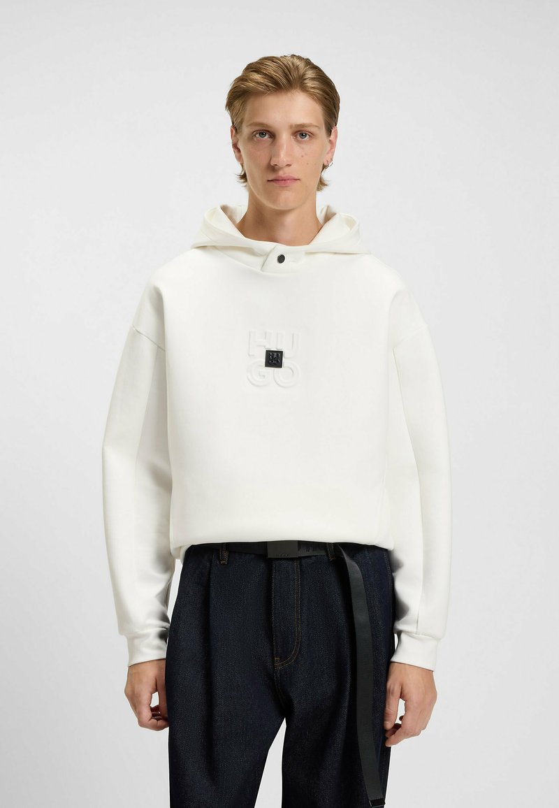 Hugo Boss hvid hættejakke med snoretræk, prægede logo på brystet og en glat tekstur, parret med mørke denim bukser og et sort bælte.