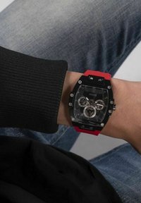 Black and red wristwatch with a rectangular case, transparent face, three sub-dials, and silver accents on a red rubber strap.