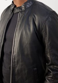 Schwarze Lederjacke mit hohem Kragen, seitlichem Reißverschluss und glatter Textur. Verfügt über minimale Nähte und eine Seitentasche.