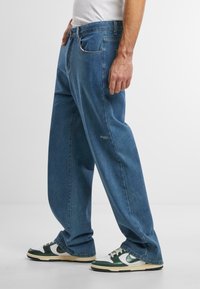 Jeans de mezclilla de color azul claro con un corte relajado, con un frente plano, bolsillos laterales y un pequeño detalle de logo en el muslo. Llevados con zapatillas verdes y blancas.