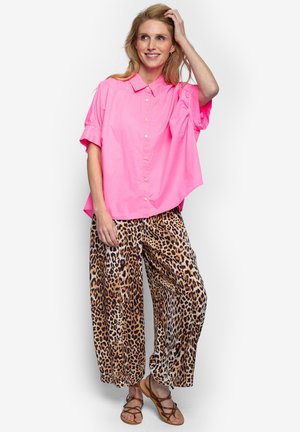 Kobieta uśmiechająca się, ubrana w jaskraworóżową koszulę na krótki rękaw z guzikami, spodnie o szerokich nogawkach z nadrukiem w panterkę oraz brązowe sandały na paski, pozująca z ręką we włosach.