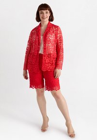 Blazer di pizzo rosso con orlo a zigzag, abbinato a pantaloni corti in pizzo rosso. Entrambi presentano motivi floreali intricati e una texture leggera.