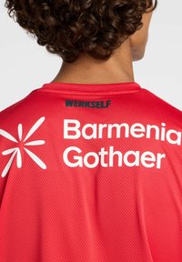 Camiseta deportiva roja con textura de malla transpirable. Presenta "WERKSELF" en negro y "Barmenia Gothaer" en impresión blanca en la parte trasera.