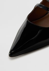 Chaussure noire en cuir verni à bout pointu avec une fine bride sur le dessus et une doublure intérieure couleur beige.