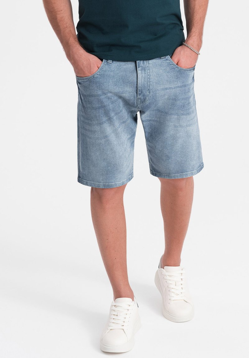 Pantalones cortos de mezclilla azul claro con corte recto, bolsillos frontales y bordes dobladillados, combinados con zapatillas blancas y una camiseta verde oscuro.