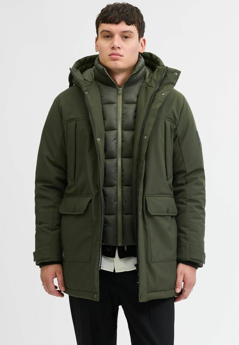 Groene geïsoleerde parka met een ritssluiting aan de voorkant, twee grote voorzakken en een afneembare capuchon, gelaagd over een gewatteerde groene binnenjas.