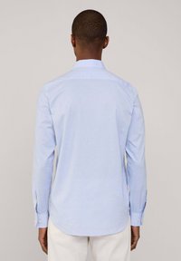 Camisa de manga larga azul claro con cuello puntiagudo, confeccionada en una tela suave. Presenta puños abotonados y un dobladillo redondeado en la parte de atrás.