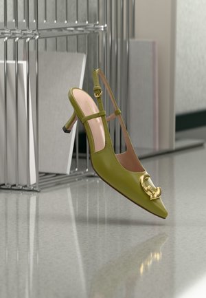 Pantof cu toc cui pentru femei, culoare verde măsliniu, cu baretă în spate și detaliu de cataramă aurie, așezat lângă un suport metalic cu obiecte de culoare neutră.