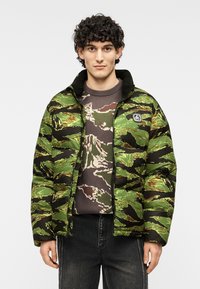 Μπουφάν τύπου puffer σε πράσινο καμουφλάζ με μαύρες λεπτομέρειες. Διαθέτει κλείσιμο με φερμουάρ και ψηλό γιακά. Φοράται πάνω από ένα πουλόβερ με σχέδιο καμουφλάζ.