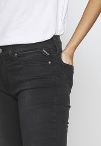 Jeans de denim negro con un ajuste slim, bolsillos frontales y una pequeña etiqueta con el logo en la cinturilla. La tela tiene una textura suave y un ligero brillo.