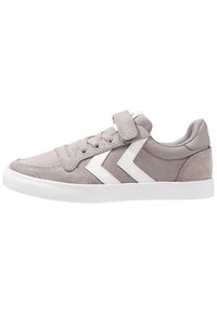 Zapatilla de deporte gris claro con rayas blancas en forma de chevrón, cierre de cordones en la parte delantera y una correa de Velcro en la parte superior, con una suela de goma blanca.