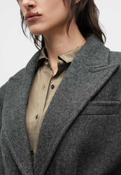 Grå uldfrakke med bred revers, der har en hakket krave og en lomme; lag på lag over en lysebrun skjorte med knapper.