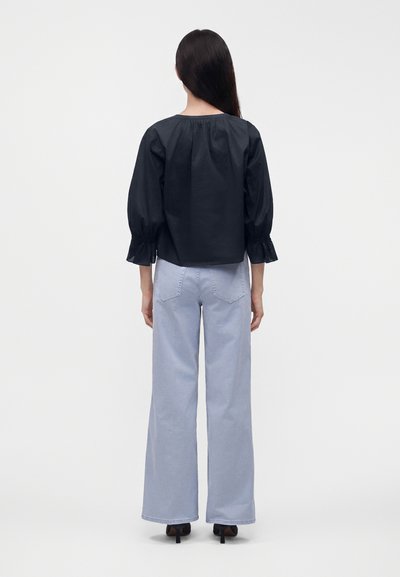 Femme aux longs cheveux foncés portant un chemisier noir à manches longues et un jean bleu clair à jambes larges, debout, dos à un fond uni.