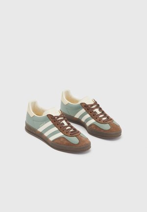 Para tenisówek Adidas Gazelle w jasnoniebieskim, brązowym i białym kolorze z brązowymi sznurowadłami na jednolitym jasnym tle.
