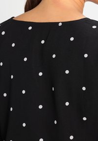 Top in tessuto nero con scollatura ampia e un motivo di pois bianchi sparsi. La texture sembra morbida e liscia.