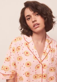 Cămașă de pijama roz cu mâneci scurte și garnitură roșie, având un model cu forme galbene de plăcinte și inimioare roșii mici. Textură moale a țesăturii.
