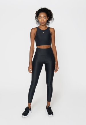 Schlanke Frau mit lockigem Haar, die ein schwarzes sportliches Crop-Top, hoch taillierte Leggings und schwarze Turnschuhe vor weißem Hintergrund trägt.