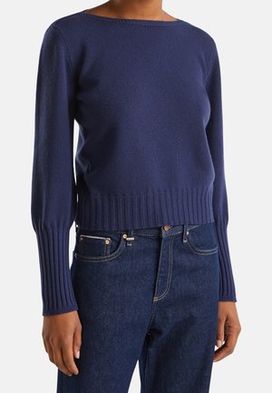 Femme portant un pull en maille côtelée bleu marine à manches longues associé à un jean en denim bleu foncé, debout devant un fond uni.
