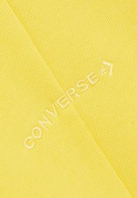Tessuto giallo con una trama a coste che presenta il logo "CONVERSE" ricamato in bianco. Il design è semplice e pulito, enfatizzando il colore.