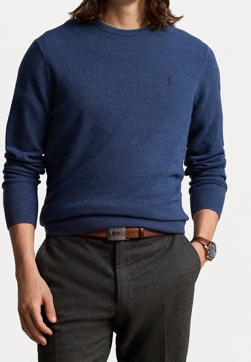 Homme portant un pull à col rond bleu, une ceinture en cuir marron, un pantalon gris et une montre-bracelet en cuir marron avec un cadran foncé.
