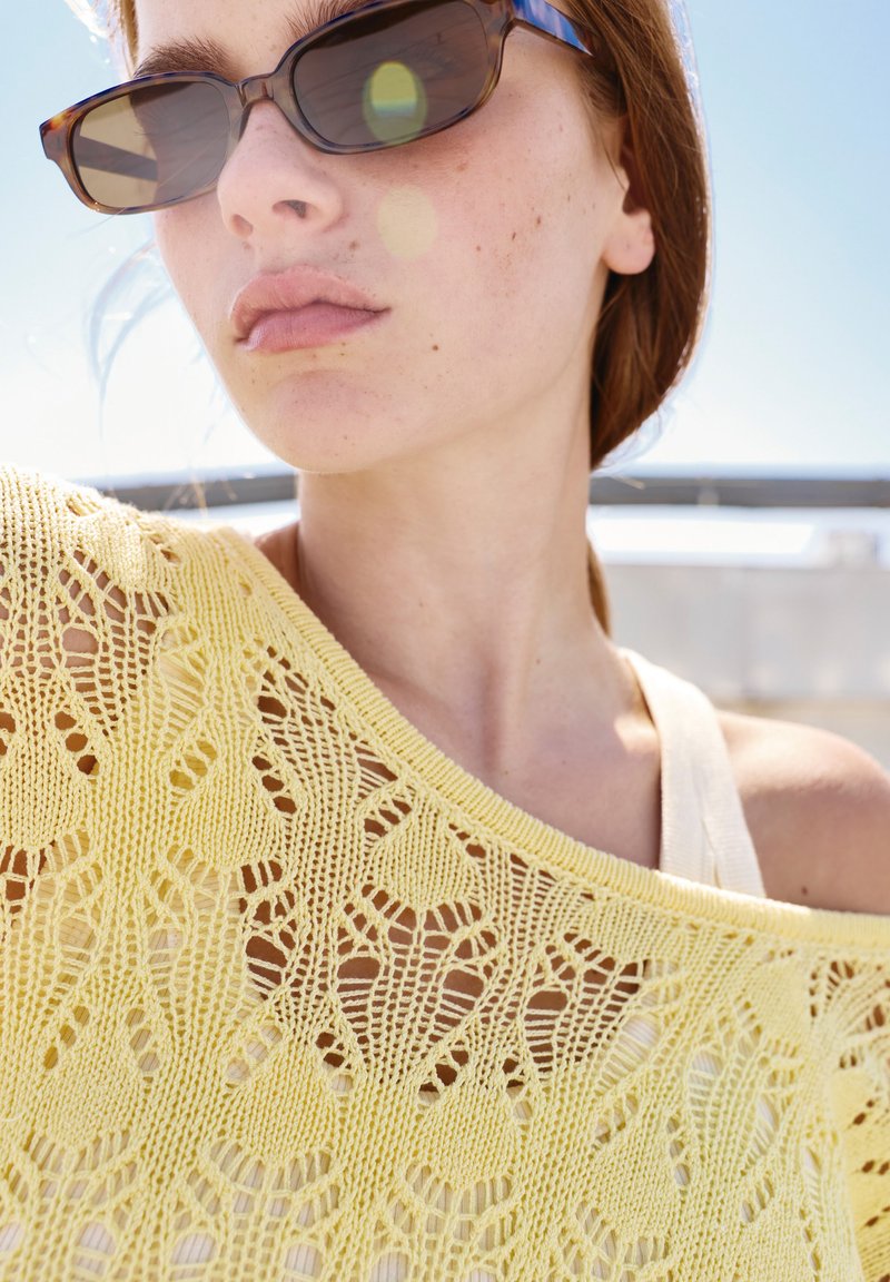 Jeune femme portant des lunettes de soleil en écaille et un haut jaune en tricot avec une bretelle blanche visible sur un ciel ensoleillé.