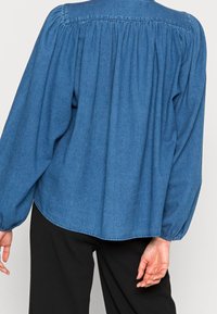 Camicia in denim con schiena arricciata, maniche a palloncino e orlo arrotondato in blu scuro, abbinata a pantaloni neri. Tessuto liscio e vestibilità casual.