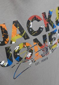 Šedé tričko s barevným flekovým potiskem a textem "Jack & Jones Essentials" v různých barvách, s hladkým textilním povrchem.