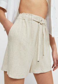 Vrouw in lichtbeige linnen shorts met een paperbag taille en strikkoord, gecombineerd met een witte cropped button-up shirt en een kant BH.